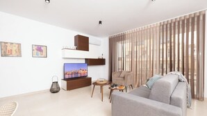 A modern living room with a grey sofa, a TV on a wooden stand, a chair, and a small table with a tray. - Modern 2-bedroom apartment in central Mellieħa. Sleeps 6 with sofa bed, A/C, Wi-Fi, Smart TV & lift access. Steps from shops, restaurants & public transport. (Il-Mellieħa)