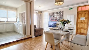 A modern kitchen with white cabinets, a dining area with a table set for two, and a living area with a sofa and a TV.