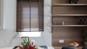 Eine moderne Küche mit Fenster und Jalousien, Holzregalen mit Pflanzen und einer Obstschale auf der Arbeitsfläche.