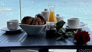 Une table de petit-déjeuner avec des croissants, un verre de jus d’orange, une tasse, un pot de confiture et une rose près d’une fenêtre donnant sur la mer.