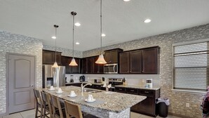 A modern kitchen with granite countertops, dark wooden cabinets, a central island with bar stools, stainless steel appliances, and pendant lighting. - Themed 8BR | Arcade | 2 Theaters | Pool & Spa (Kissimmee)