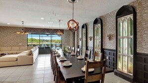 A dining area with a long table set for a meal, surrounded by wooden chairs, with a stone wall and large windows offering a view of the outdoors. - Themed 8BR | Arcade | 2 Theaters | Pool & Spa (Kissimmee)