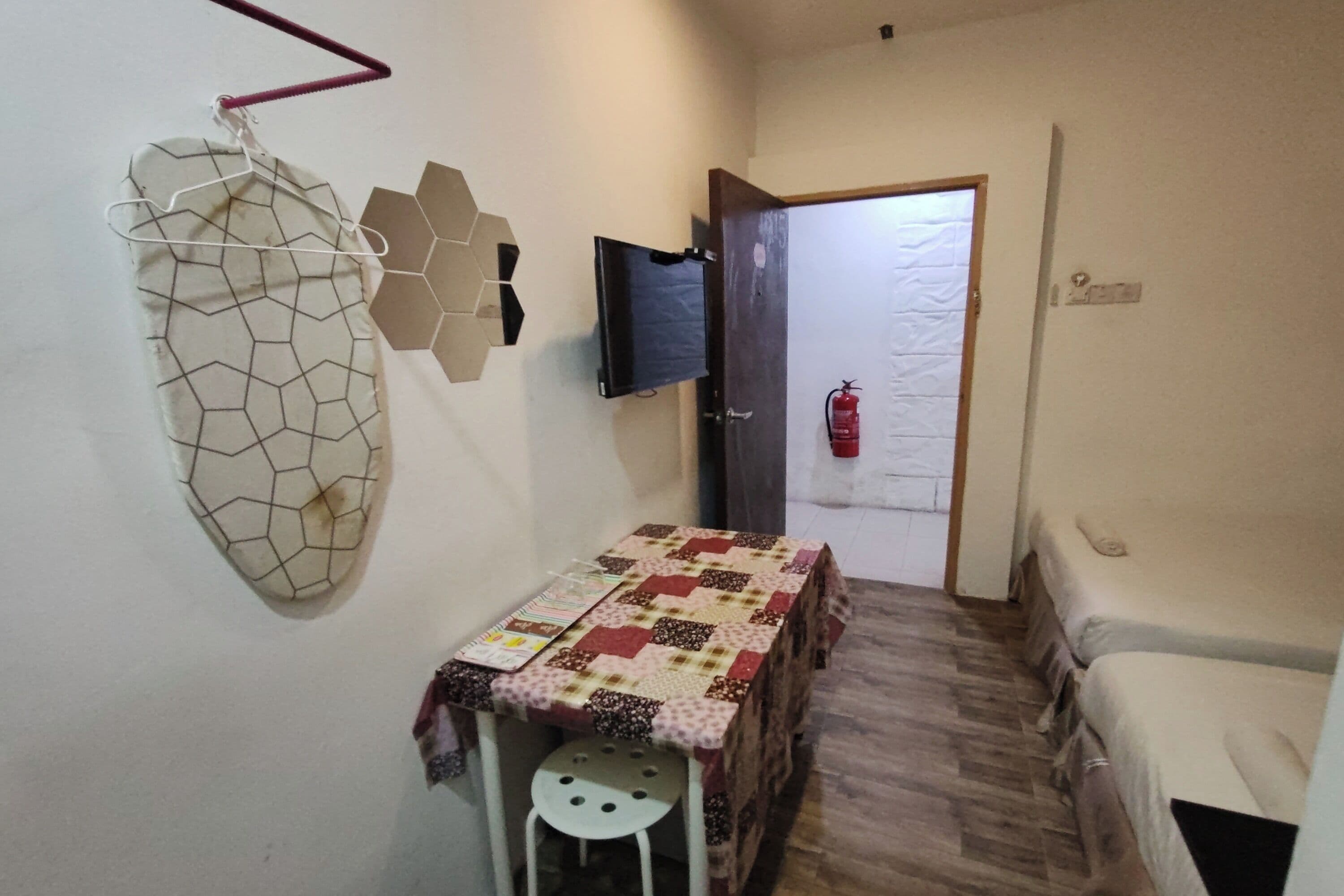 Una habitación pequeña y funcional con suelo de madera, una mesa con mantel estampado, una silla blanca, un televisor montado en la pared y una puerta con extintor.