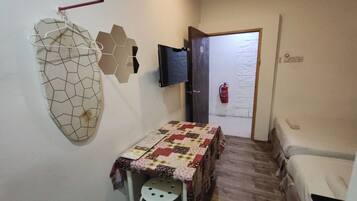 Una habitación pequeña y funcional con suelo de madera, una mesa con mantel estampado, una silla blanca, un televisor montado en la pared y una puerta con extintor.