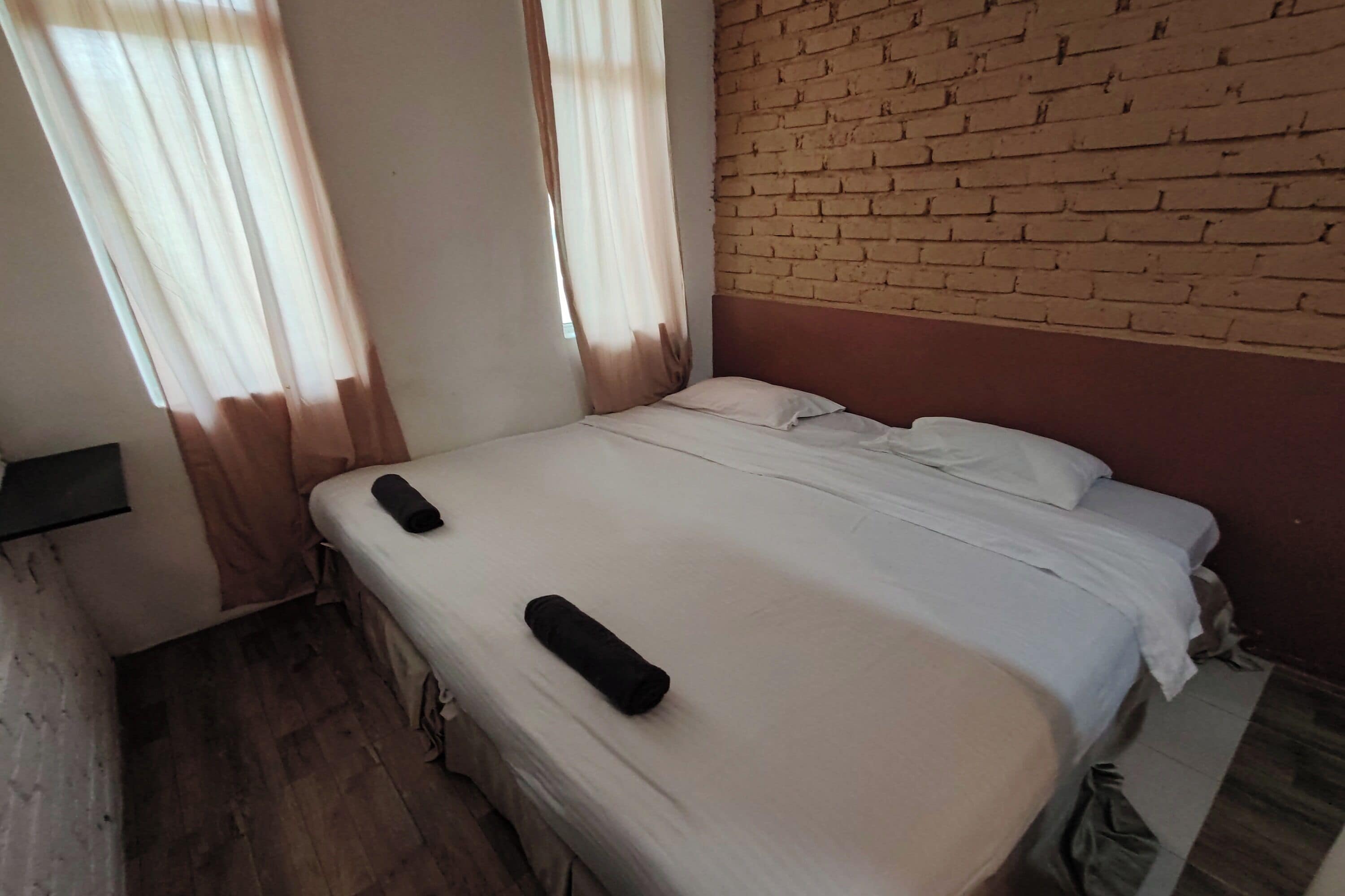 Uma cama de solteiro com colchão branco e duas almofadas, piso de madeira, parede de tijolos e janela com cortinas.