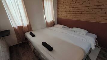 Uma cama de solteiro com colchão branco e duas almofadas, piso de madeira, parede de tijolos e janela com cortinas.