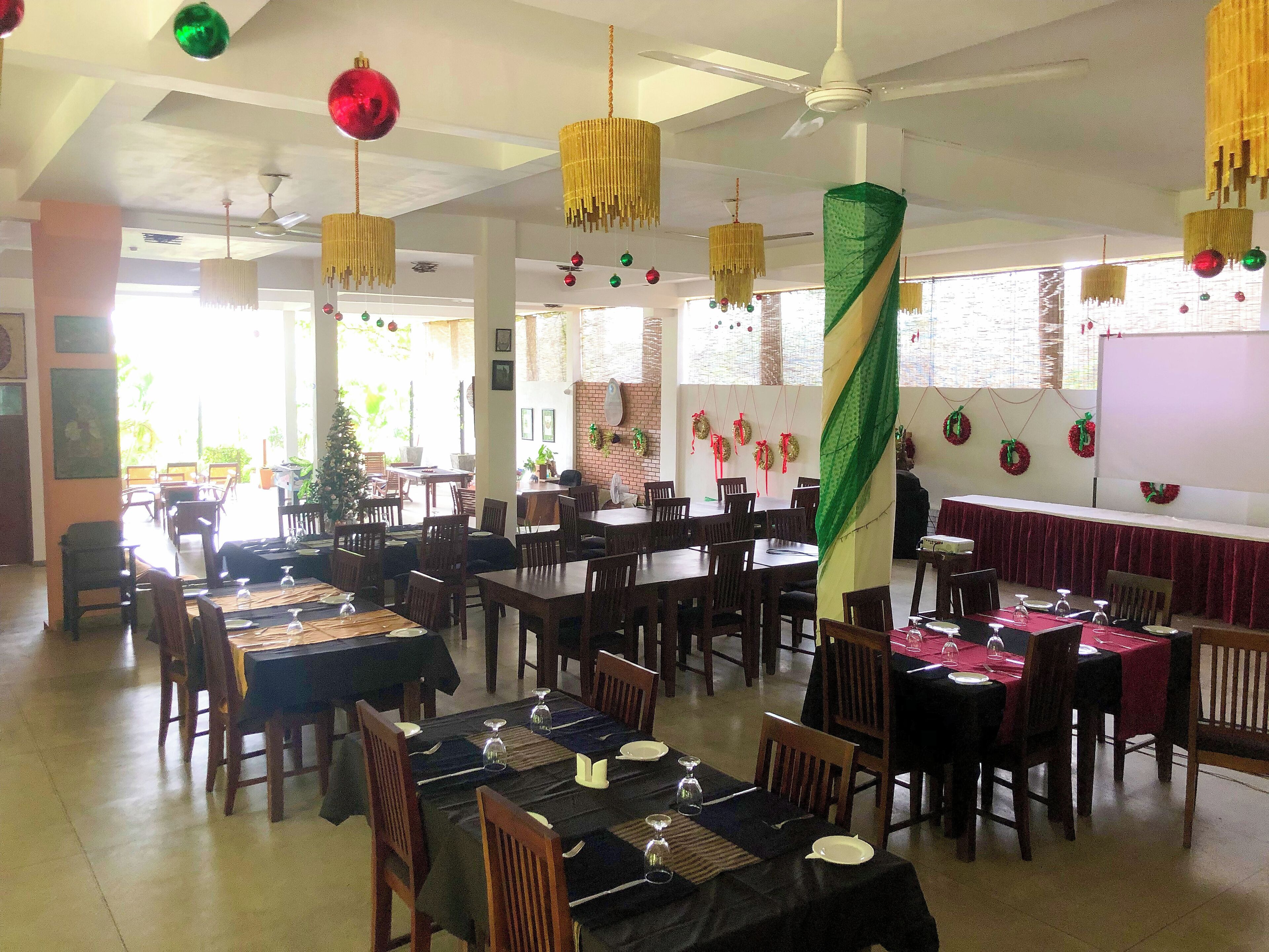 Uma sala de jantar com várias mesas preparadas para hóspedes, apresentando diferentes opções de assento e um ambiente festivo com decorações penduradas.