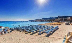 A beach with rows of blue and white striped umbrellas and sun loungers, a clear blue sky, and a calm sea. - Modern designer apartment in sunny Mellieħa Bay! Enjoy sea views, stylish comfort, and nearby beaches like Golden Bay & Comino — perfect for a relaxing family getaway (Il-Mellieħa)
