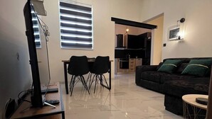 A modern living room with a black sofa, a dining area with two chairs, a kitchen in the background, and a ceiling with geometric pendant lights.