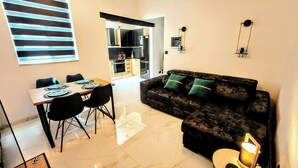 A modern living room with a dark-colored sofa, a dining table with chairs, and a kitchen visible through an open doorway.
