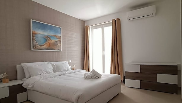 A modern bedroom with a bed, nightstands, a painting, a window with curtains, and an air conditioning unit. - This apartment is a 2 bedroom(s), 2 bathrooms, located in Il-Mellieħa, null. (Il-Mellieħa)