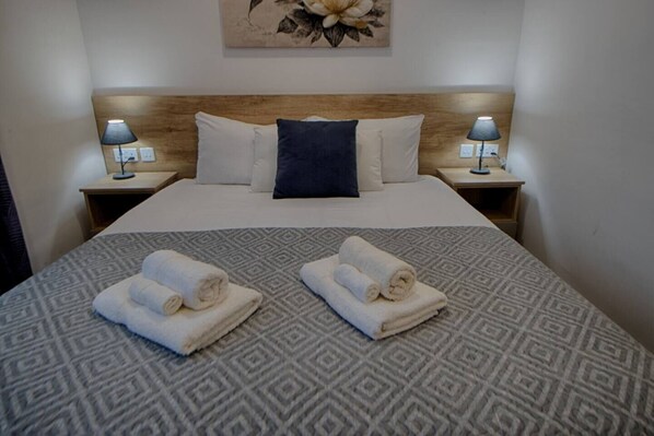 Una cama bien tendida con una colcha estampada, dos toallas enrolladas sobre la cama y un cabecero de madera.
