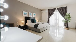 A modern hotel room with a large bed, bedside lamps, a vanity with lights, a potted plant, and a large window with curtains. - Escape to your dream coastal retreat in Mellieħa — a spacious, modern haven just steps from golden beaches. Relax, unwind, and soak up the island sunshine. (Il-Mellieħa)