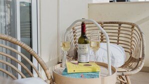 A wicker chair with a small round table on a balcony. The table has a bottle of wine, a glass, and a book. - Escape to your dream coastal retreat in Mellieħa — a spacious, modern haven just steps from golden beaches. Relax, unwind, and soak up the island sunshine. (Il-Mellieħa)