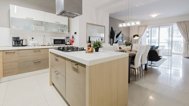 A modern kitchen with a central island, a dining area with a black chair, and a large window with curtains. - Escape to your dream coastal retreat in Mellieħa — a spacious, modern haven just steps from golden beaches. Relax, unwind, and soak up the island sunshine. (Il-Mellieħa)
