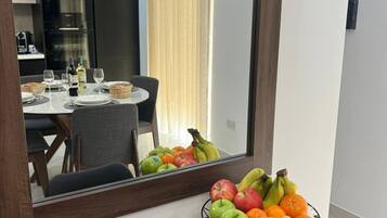 A modern room with a dining area visible in the mirror reflection, a fruit bowl on the counter, and a kitchen with appliances in the background.