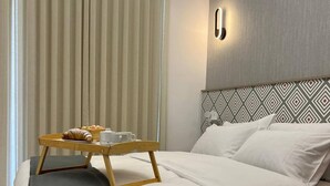 Una habitación de hotel moderna con una cama impecable, una mesita de noche con una bandeja de desayuno, y un ventanal grande con persianas verticales.