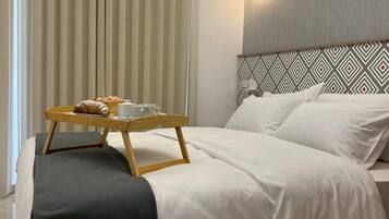 Ein modernes Hotelzimmer mit einem ordentlich bezogenen Bett, einem Nachttisch mit einem Tablett Frühstücksangeboten und einem großen Fenster mit vertikalen Jalousien.