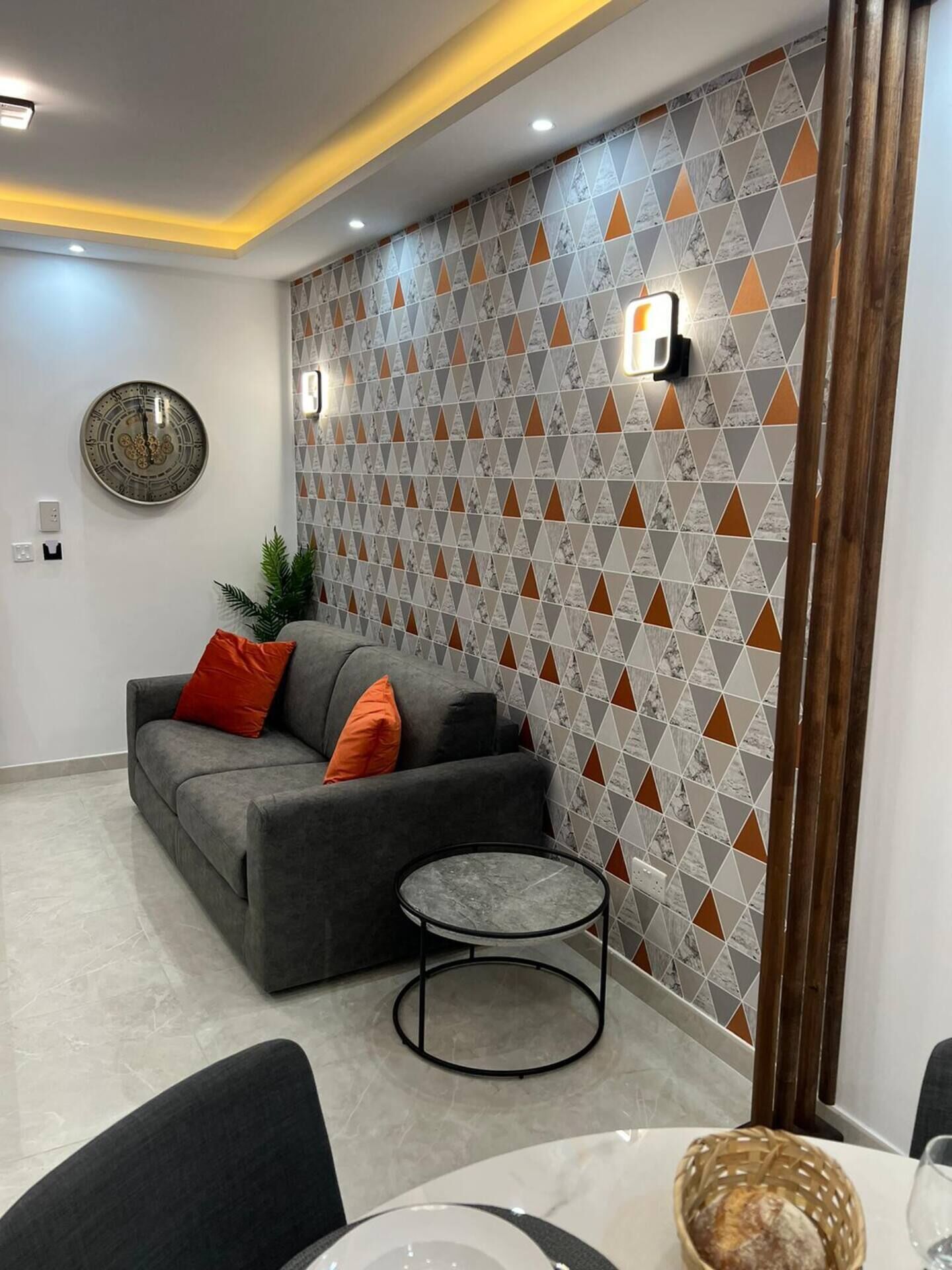Ein modernes Wohnzimmer mit einem grauen Sofa, orangefarbenen Kissen, einem runden Couchtisch und einer Wand mit Muster.