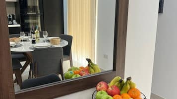 Quarto moderno com área de refeições visível no reflexo do espelho, uma tigela de frutas na bancada e cozinha com eletrodomésticos ao fundo.