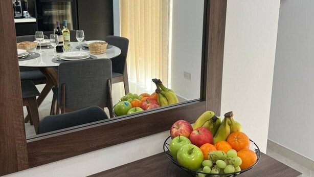 Een moderne kamer, met in de spiegel een eethoek te zien, een schaal fruit op het aanrecht en een keuken met apparatuur op de achtergrond.