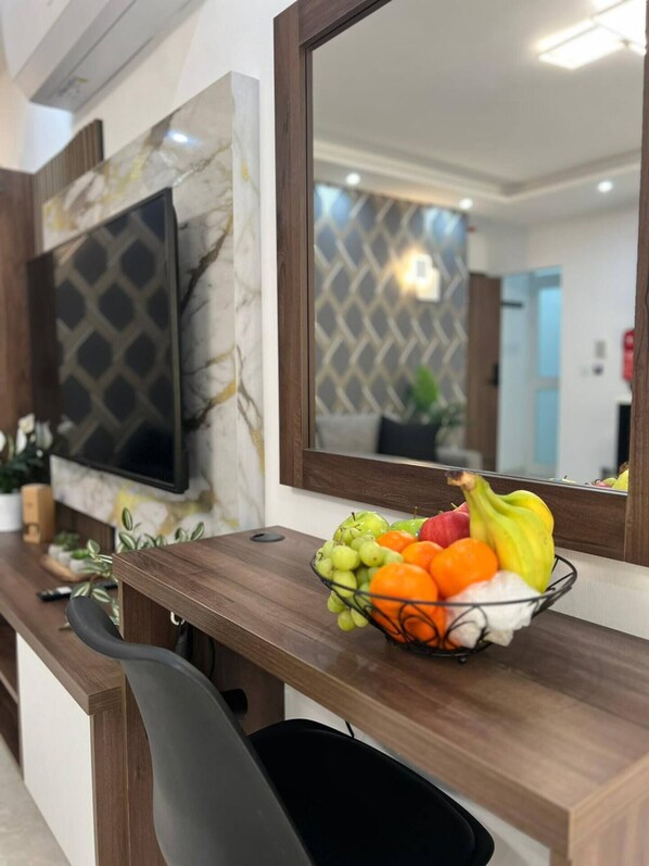 A modern living room with a TV mounted on a marble wall, a wooden console table with a fruit bowl, and a large mirror reflecting the room.