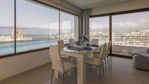 A modern dining room with a glass table and chairs, large windows with a city view, and a balcony overlooking a body of water.