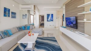 A modern living room with a sofa, coffee table, and TV. There are paintings on the walls and a window with curtains.