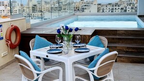 A rooftop dining area with a table set for two, overlooking a cityscape and a swimming pool.