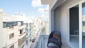 Un balcón con una silla de mimbre, vista a una calle de la ciudad y un edificio con una grúa al fondo.