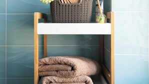 Un baño con pared de azulejos azules, un estante de madera con una cesta que contiene toallas y una flor, y una silla de madera.