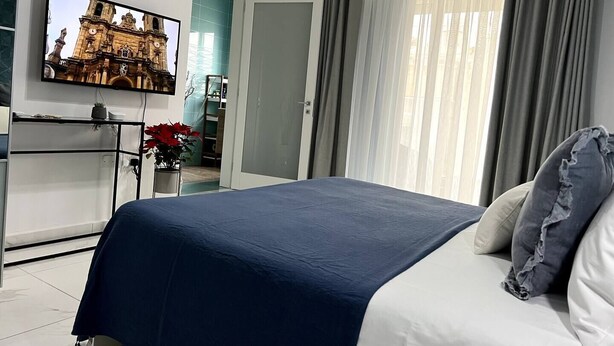 Un dormitorio con cama, televisor montado en la pared, planta en maceta y vista a un edificio a través de la ventana.