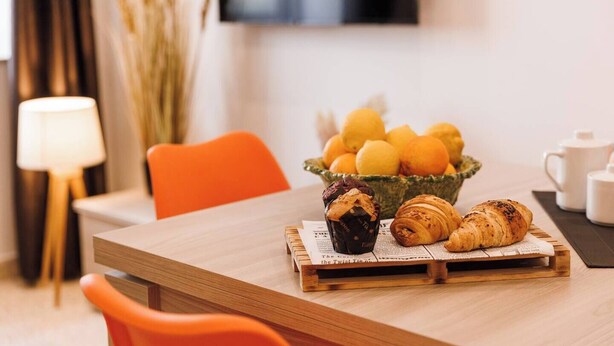 A dining area with a wooden table, a bowl of oranges, a croissant, and a muffin.