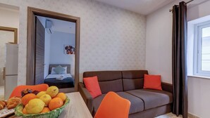 A compact room with a bed, a brown sofa with orange cushions, a wooden table with a fruit bowl, and a window with curtains.