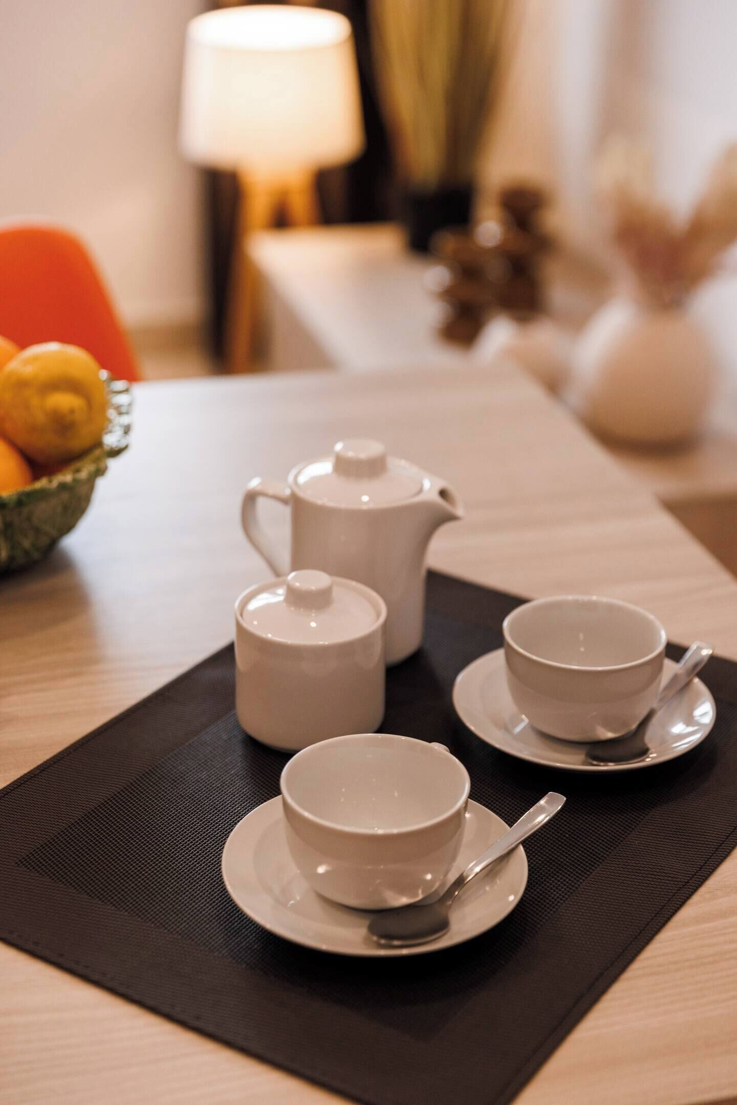 A neatly arranged tea set on a black placemat, including a teapot, two cups, and a saucer, with a bowl of fruit and a lamp in the background.