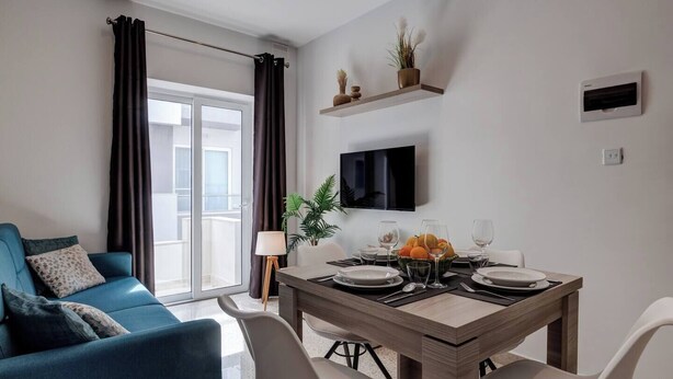 A modern living room with a blue sofa, a dining table set for four, and a TV mounted on the wall. - This apartment is a 2 bedroom(s), 1 bathrooms, located in Il-Mellieħa, null. (Il-Mellieħa)