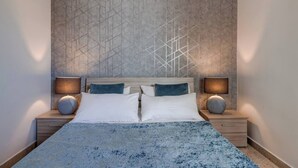 A modern bedroom with a geometric patterned wall, a large bed with white and blue bedding, and two bedside lamps. - This apartment is a 2 bedroom(s), 1 bathrooms, located in Il-Mellieħa, null. (Il-Mellieħa)