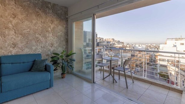 Ein modernes Wohnzimmer mit einem blauen Sofa, einem Topf mit Pflanze und einem Balkon mit Stadtblick.