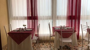 Un coin repas avec deux tables, chacune recouverte d’une nappe rouge et entourée de chaises blanches. La pièce est dotée d’un sol carrelé et de grandes fenêtres ornées de rideaux blancs légers et de draperies rouges.