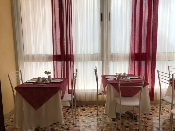 A dining area with two tables, each with a red tablecloth and white chairs. The room has a tiled floor and large windows with sheer white curtains and red drapes. - B&B Myosotis (Pisa)