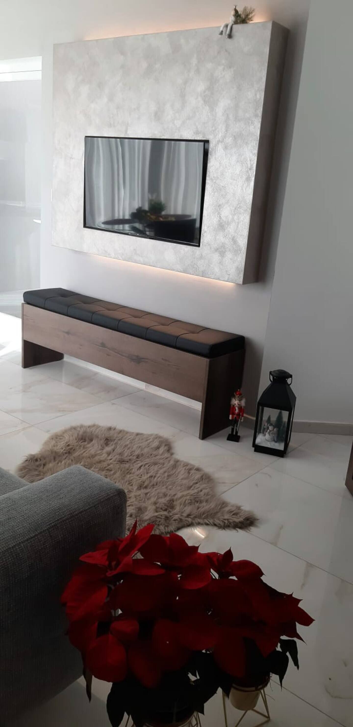 Een moderne woonkamer met een grote flatscreen-tv aan een gestructureerde wand, een houten bankje, een pluizig vloerkleed, een potplant en een decoratieve lantaarn.
