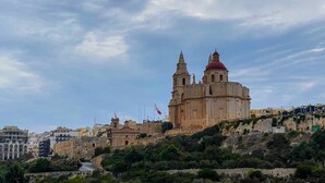 A historic building with two prominent towers and a red dome, situated on a hillside with surrounding urban structures. - This apartment is a 2 bedroom(s), 2 bathrooms, located in Il-Mellieħa, null. (Il-Mellieħa)