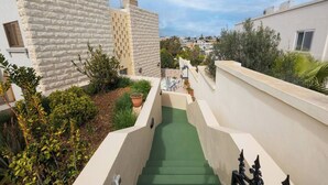 Ein schmaler, grün gefliester Weg führt zu einem Gebäude mit einer Steinaußenfassade und einem kleinen Gartenbereich mit Blumentöpfen.