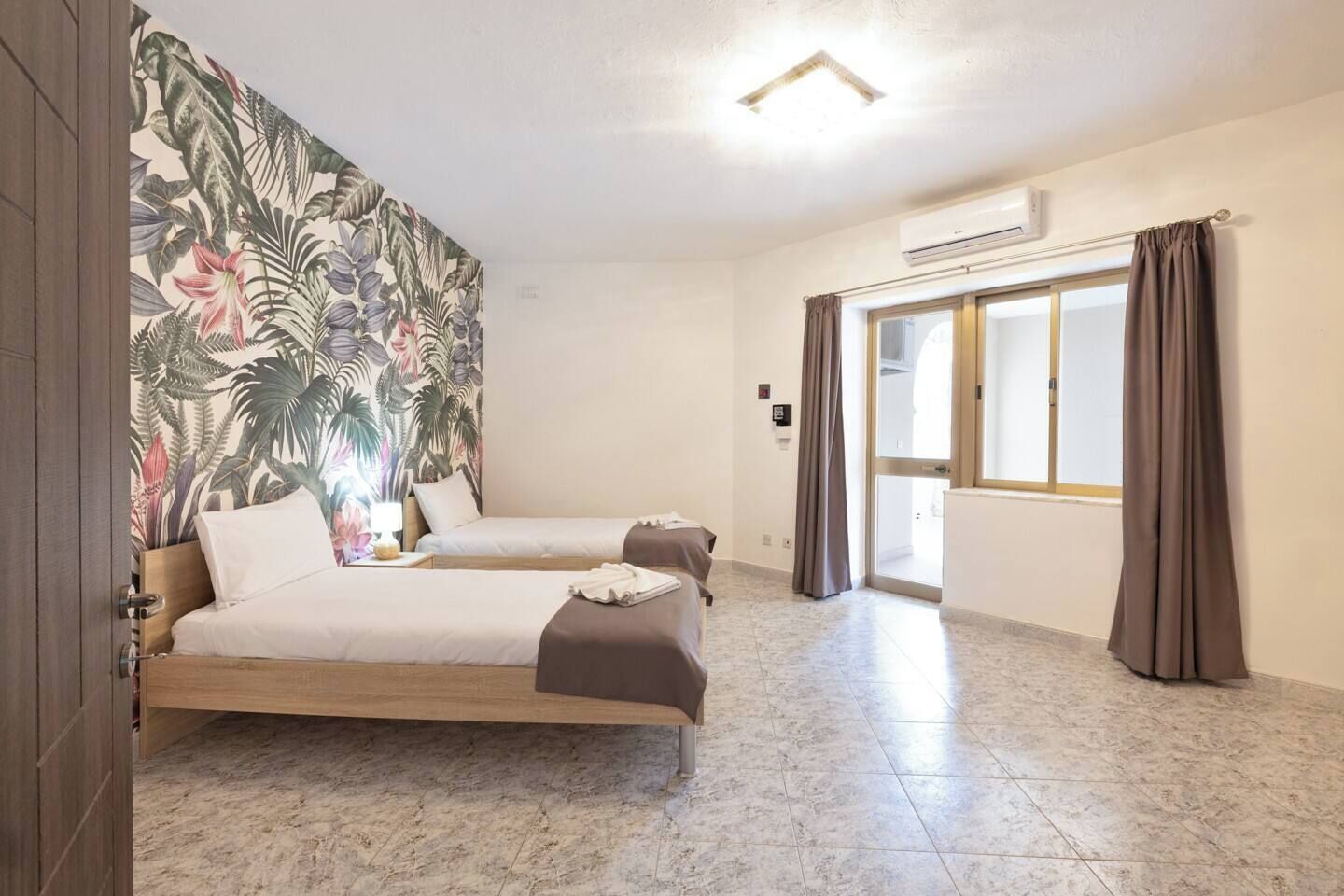 A hotel room with two beds, a tropical wallpaper, a large window with curtains, and a tiled floor.
