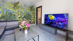 Ein Wohnzimmer mit einem großen Fernseher, einer Couch, einem Couchtisch mit Blumen und einer Wandmalerei von tropischen Blättern.
