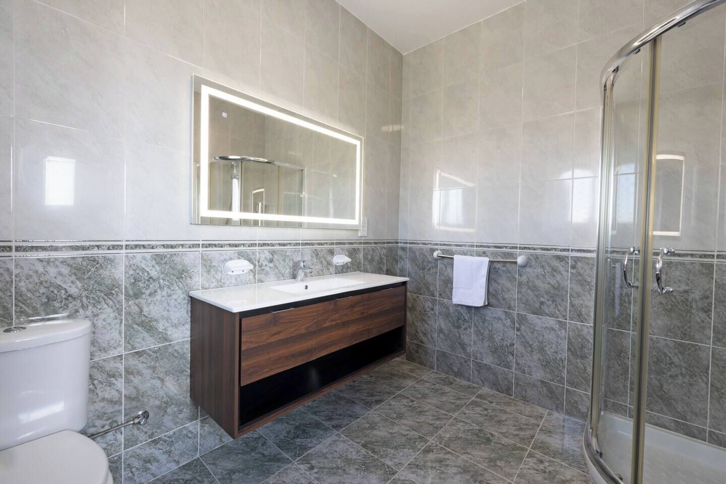 Un baño con suelo de baldosas de mármol, un tocador de madera con espejo, un inodoro y una cabina de ducha de cristal.