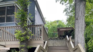 Eine Holztreppe führt zu einer Terrasse mit Geländer, umgeben von Bäumen und einem Haus.