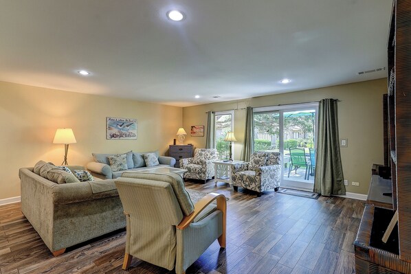 A spacious living room with a sofa set, a coffee table, and a patterned armchair. There is a sliding glass door leading to an outdoor area.