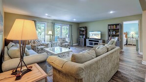 A spacious living room with a sofa, coffee table, and a television. There are bookshelves and a dining area visible. - Free 2 hours pickleball/tennis daily, pet friendly! 629 Queens Grant (Hilton Head Island)