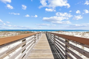 Ein hölzerner Steg führt zu einem Strand mit blauem Himmel und Wolken.